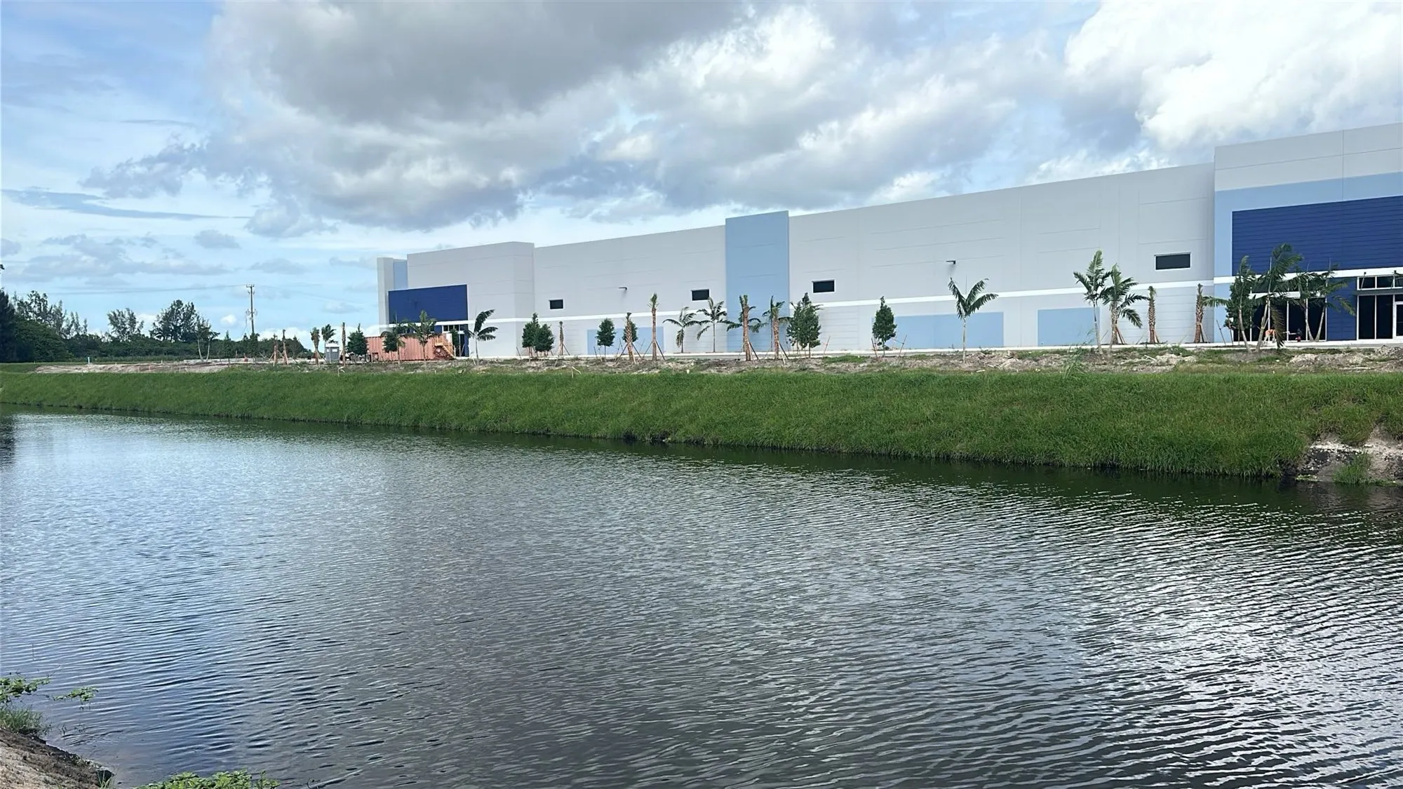 A canal with rippled water in the foreground. Its far bank is an even grassed slope; behind it stands a long white and blue commercial building with a row of newly planted palms.