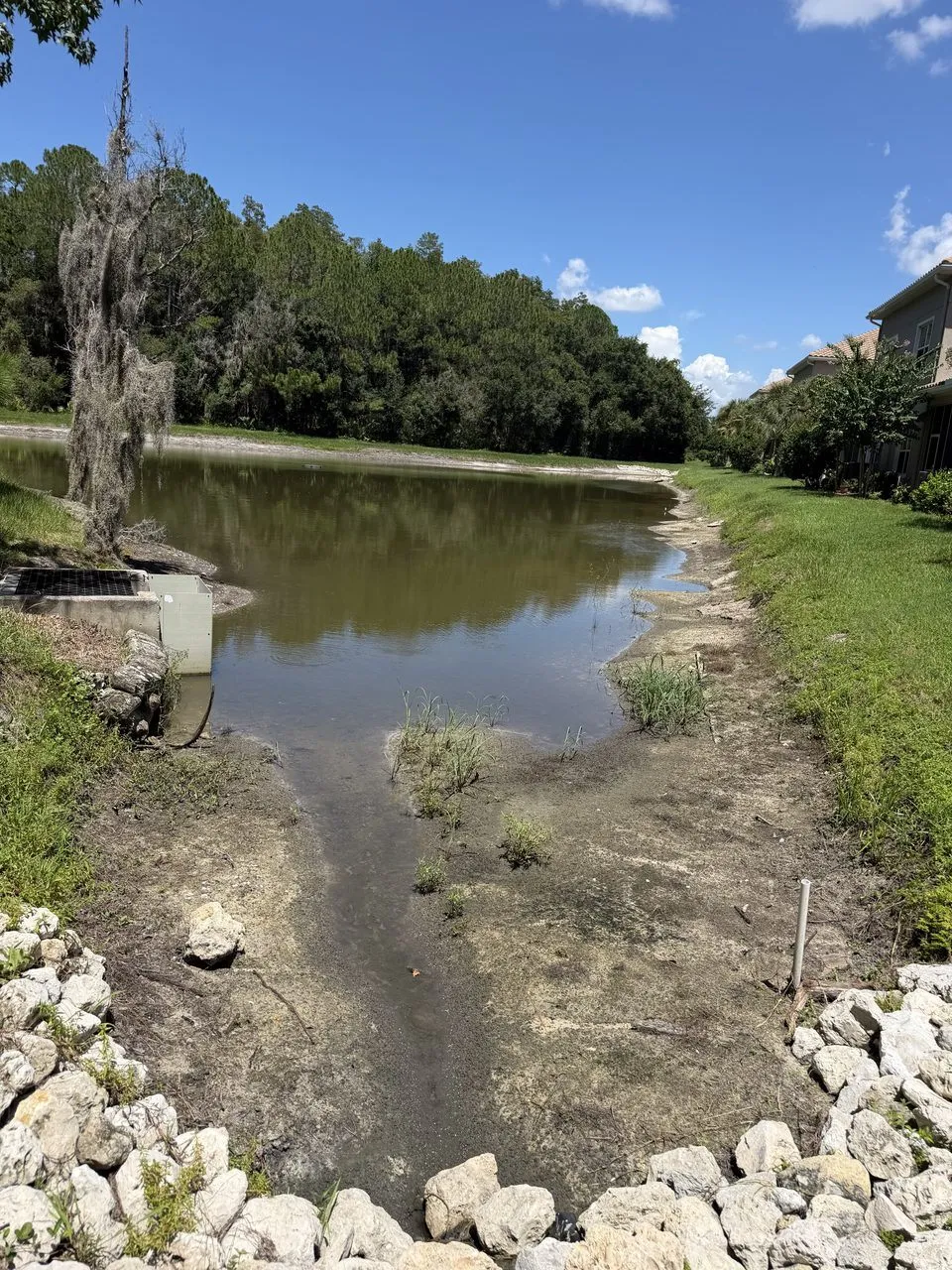 A lake at low water. A wide strip of bare, sandy lake bed with a few tufts of grass lies between a lawn and the water. A low concrete box with a grate on top stands at the edge on the left, and pale stones lie in the foreground.