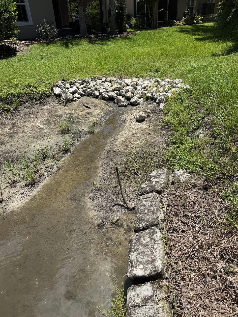 A pile of pale stones at the edge of a lawn. Below it, a narrow stream of water runs across bare, sandy soil. A row of weathered blocks lies on the right.