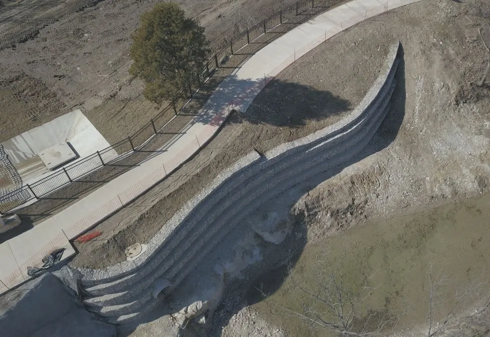 A curved retaining wall built of rock-filled wire baskets in stepped tiers, seen from above. A concrete path with a metal fence runs along the slope above it, and a pipe outlet opens at the foot of the wall.