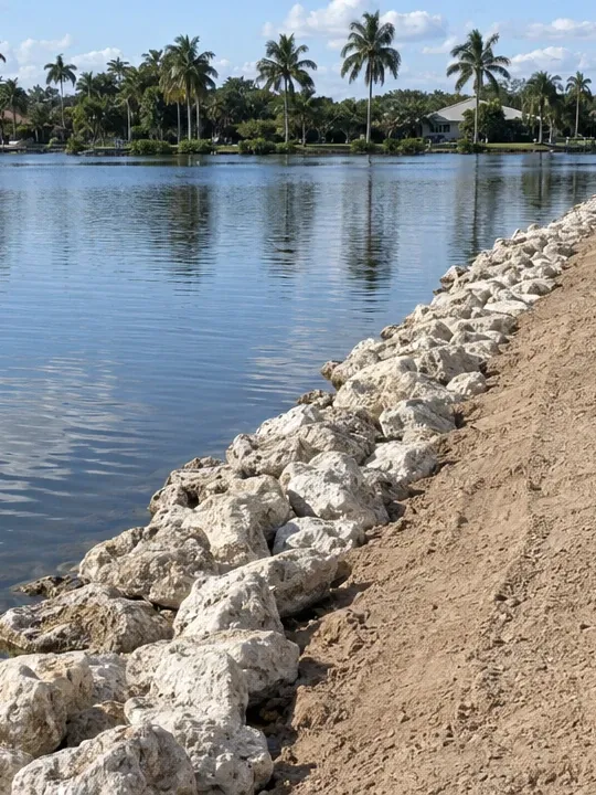 New limestone riprap along a lake shore, with a strip of bare graded sand above it and a mown lawn at the top.
