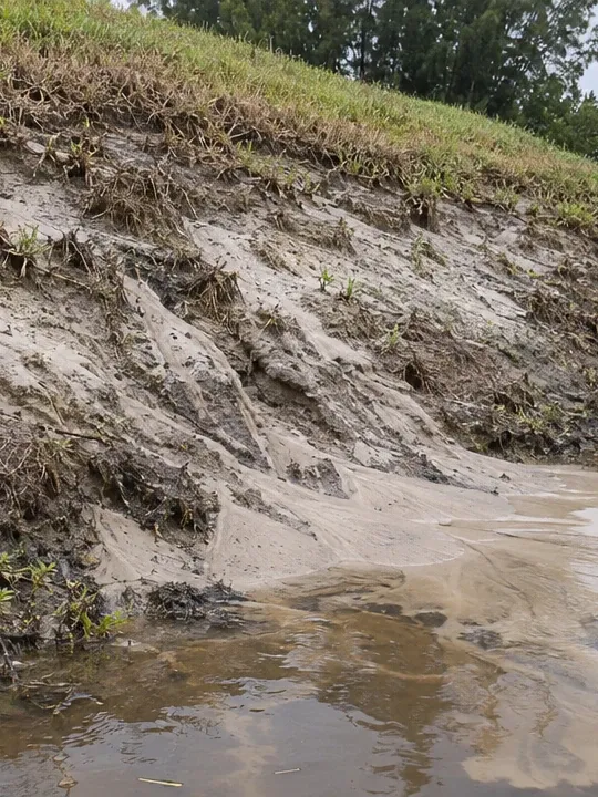 The bare side slope of a retention pond cut by erosion gullies, with sand washed into the brown water below.