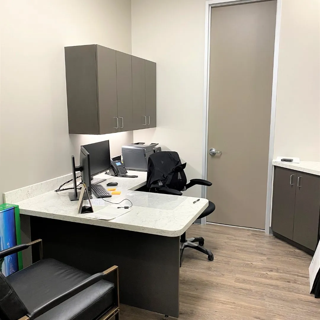 A small office: an L-shaped desk with a light stone top, two monitors, a desk telephone and a printer, gray wall cabinets above the desk, a closed door and a black office chair.