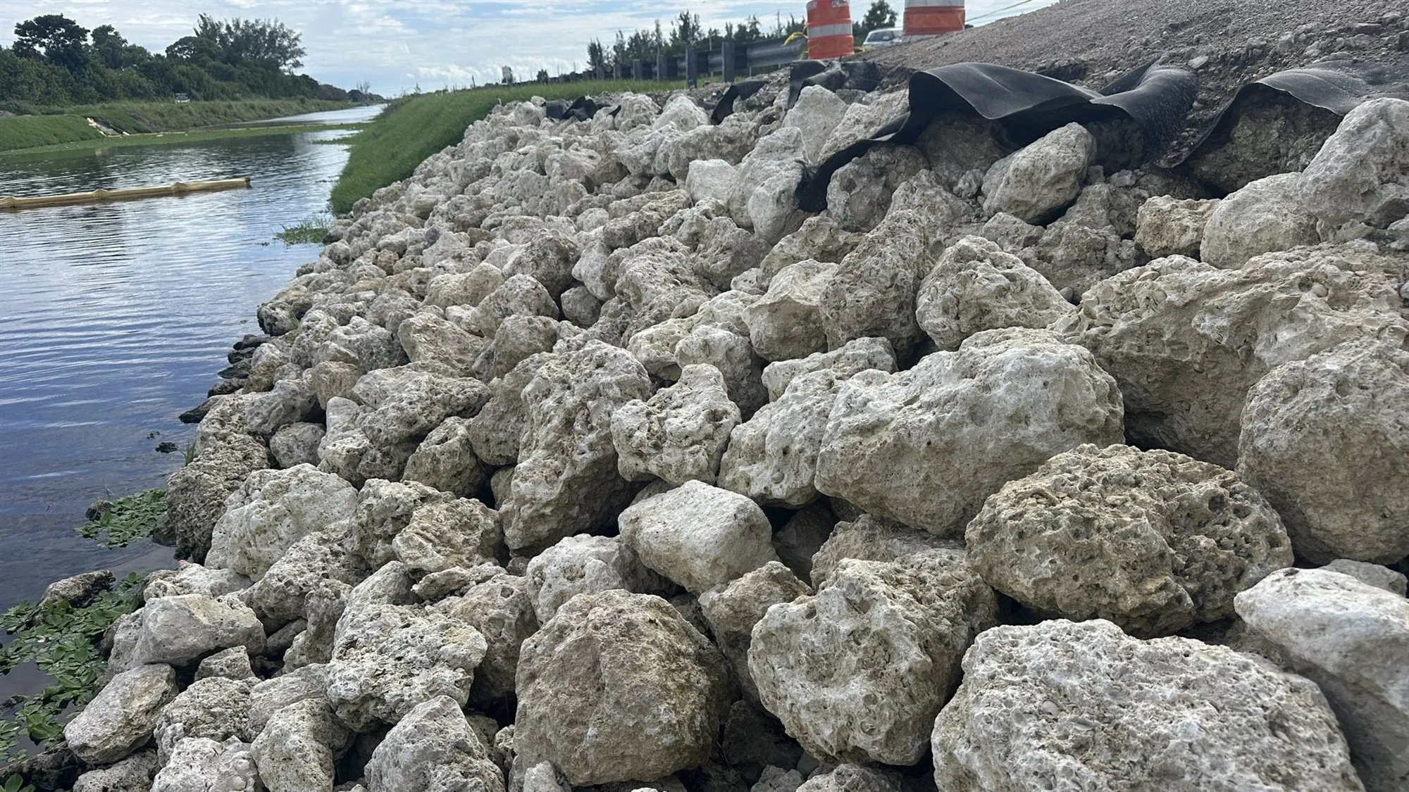 A slope of pale, rough stone seen from close up, running down to the water of a canal. At the top of the slope, sheets of black fabric stick out from under the stone.