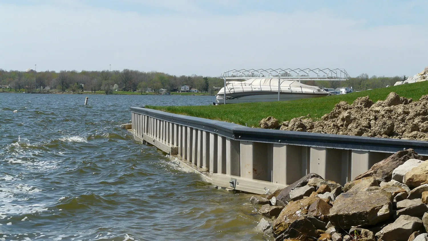 A vinyl sheet-pile wall along a lake bank, seen from one end. Gray corrugated sheets stand side by side under a dark cap, a beam runs along their face at the waterline, and large stones are piled against the end of the wall.