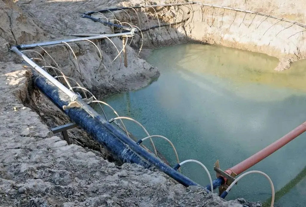 An open excavation in sandy soil with water standing in the bottom. Pipes run along its edges, and rows of thin curved hoses lead from the pipes into the ground.